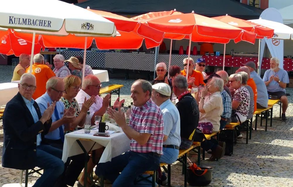 Gäste an Biertischen applaudieren unter roten Sonnenschirmen