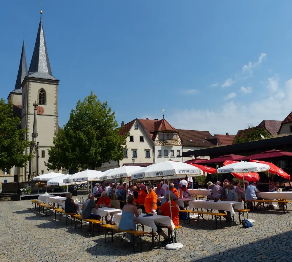 Tafel-Fest zum 20-jährigen Jubiläum auf dem Marktplatz in Haßfurt: Gäste an Biertischen unter Sonnenschirmen, im Hintergrund die Ritterkapelle